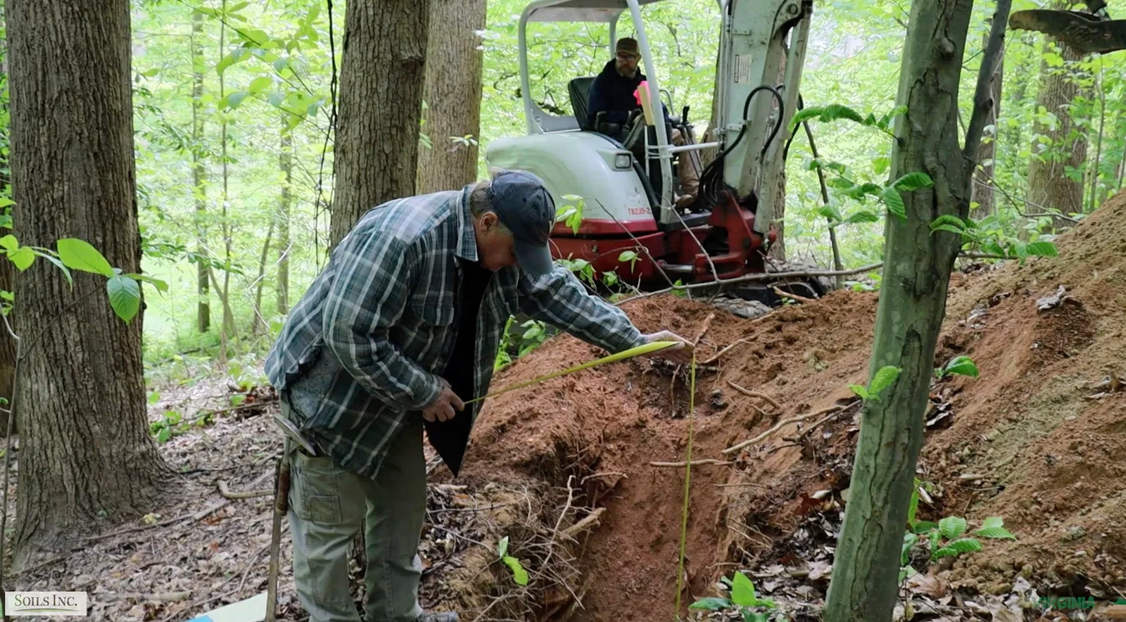Worker measuring a soil test pit beside an excavator